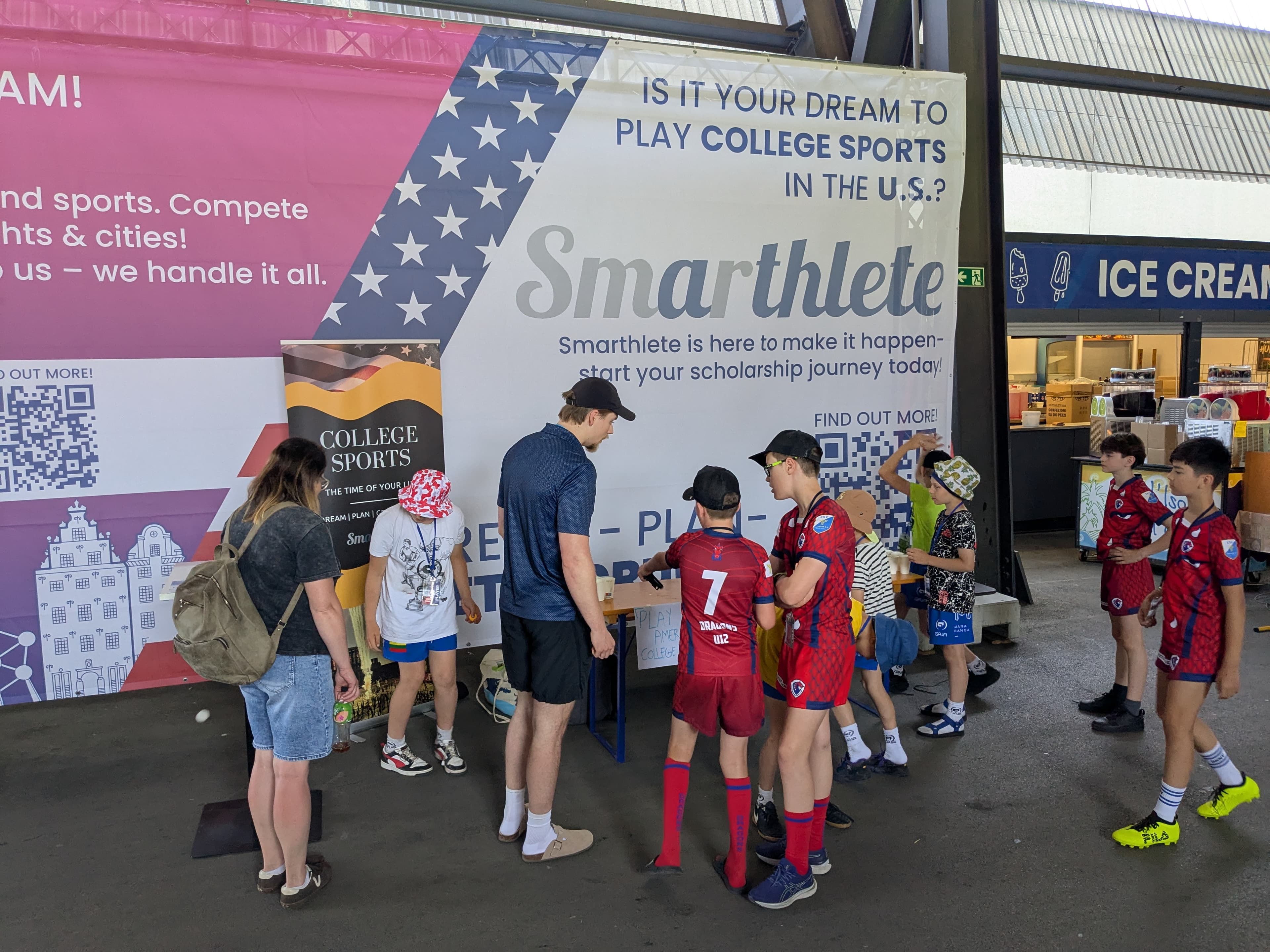 A large Smarthlete promotional banner at an information stand asking 'Is it your dream to play college sports in the U.S.?' with young athletes in front of it.