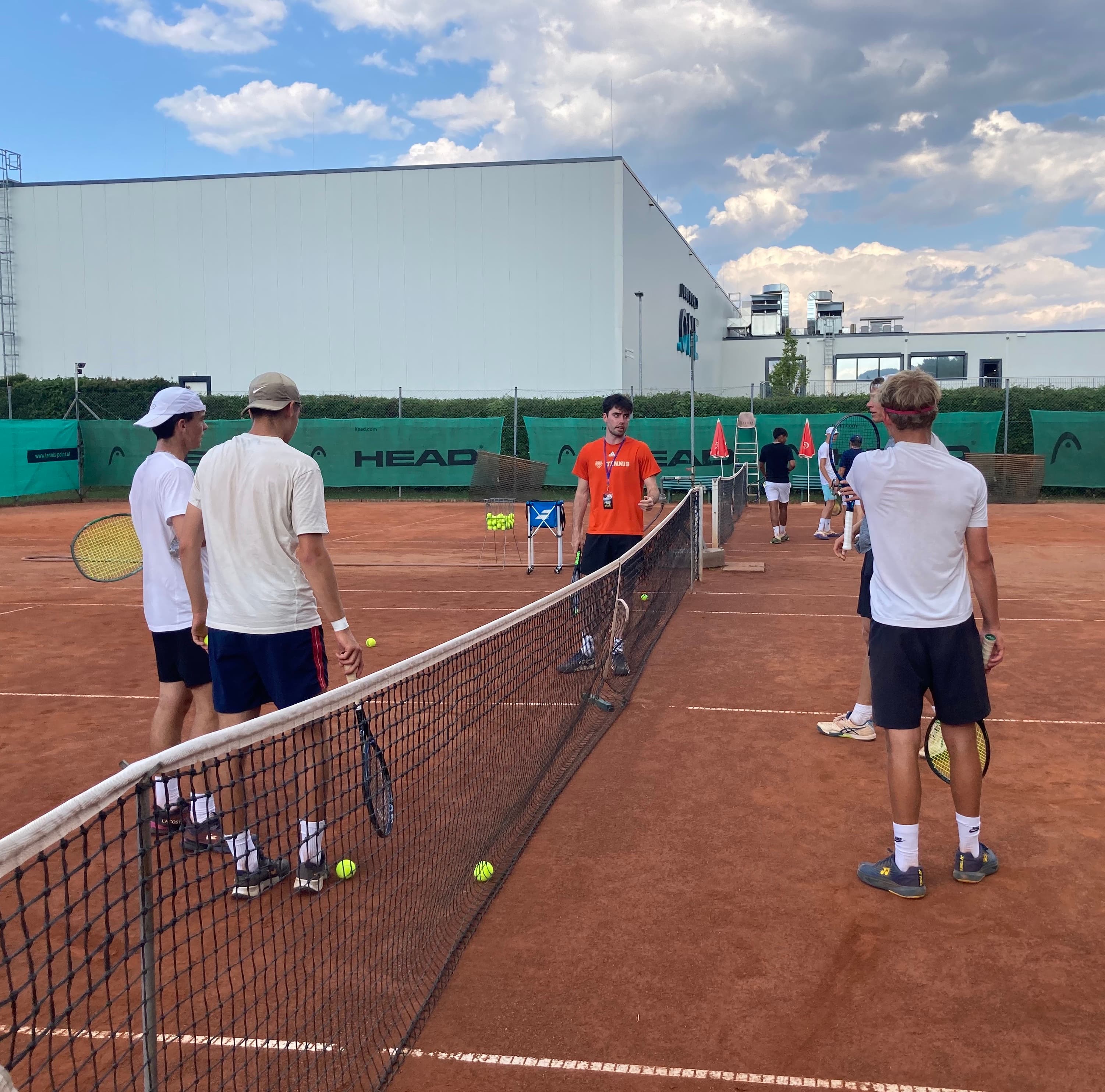 A group of tennis players standing on a red clay court during a practice session, listening to a U.S. college coach in an orange shirt.
