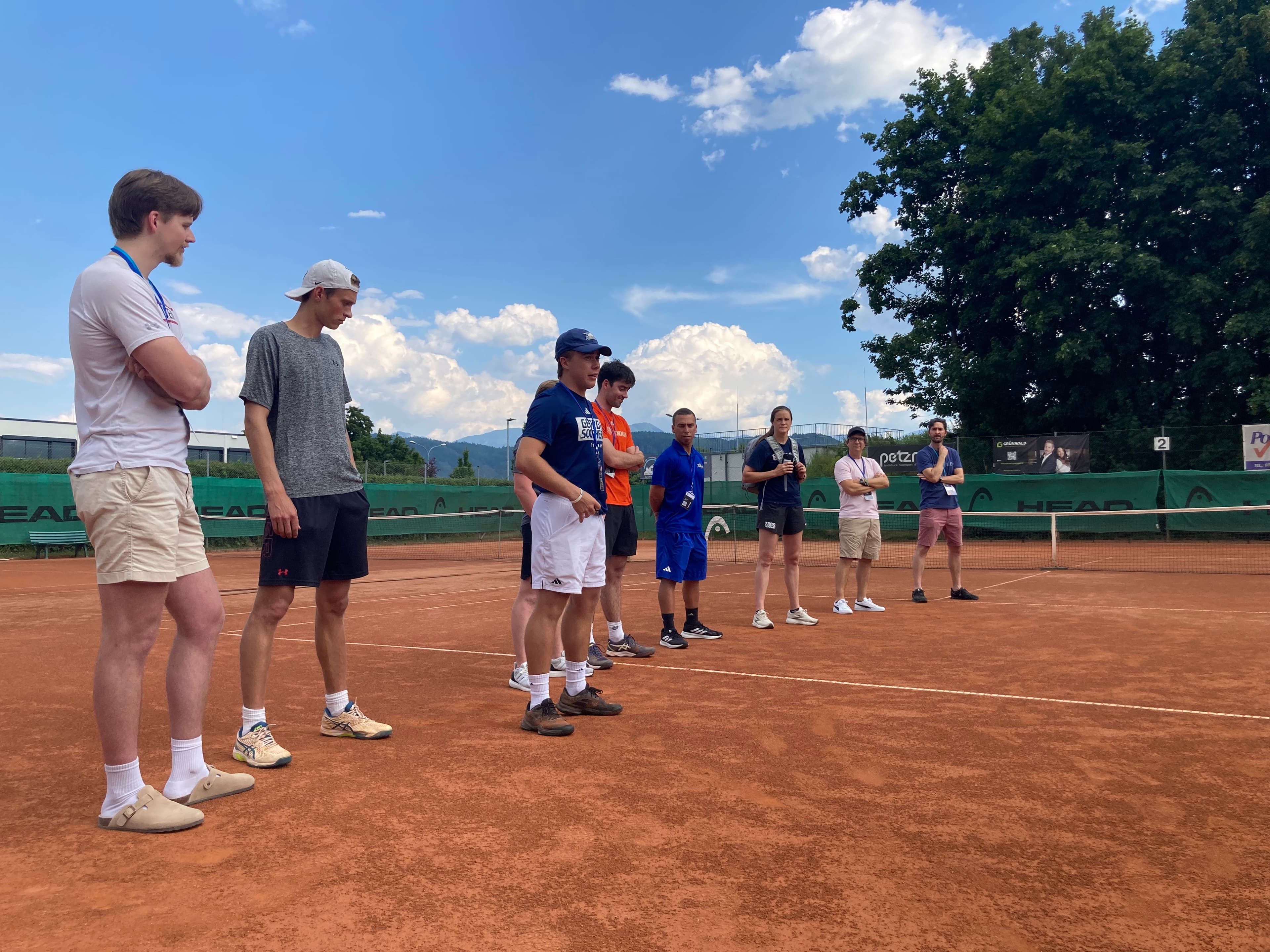 A group of college tennis coaches and college recruiting experts standing on a clay court.