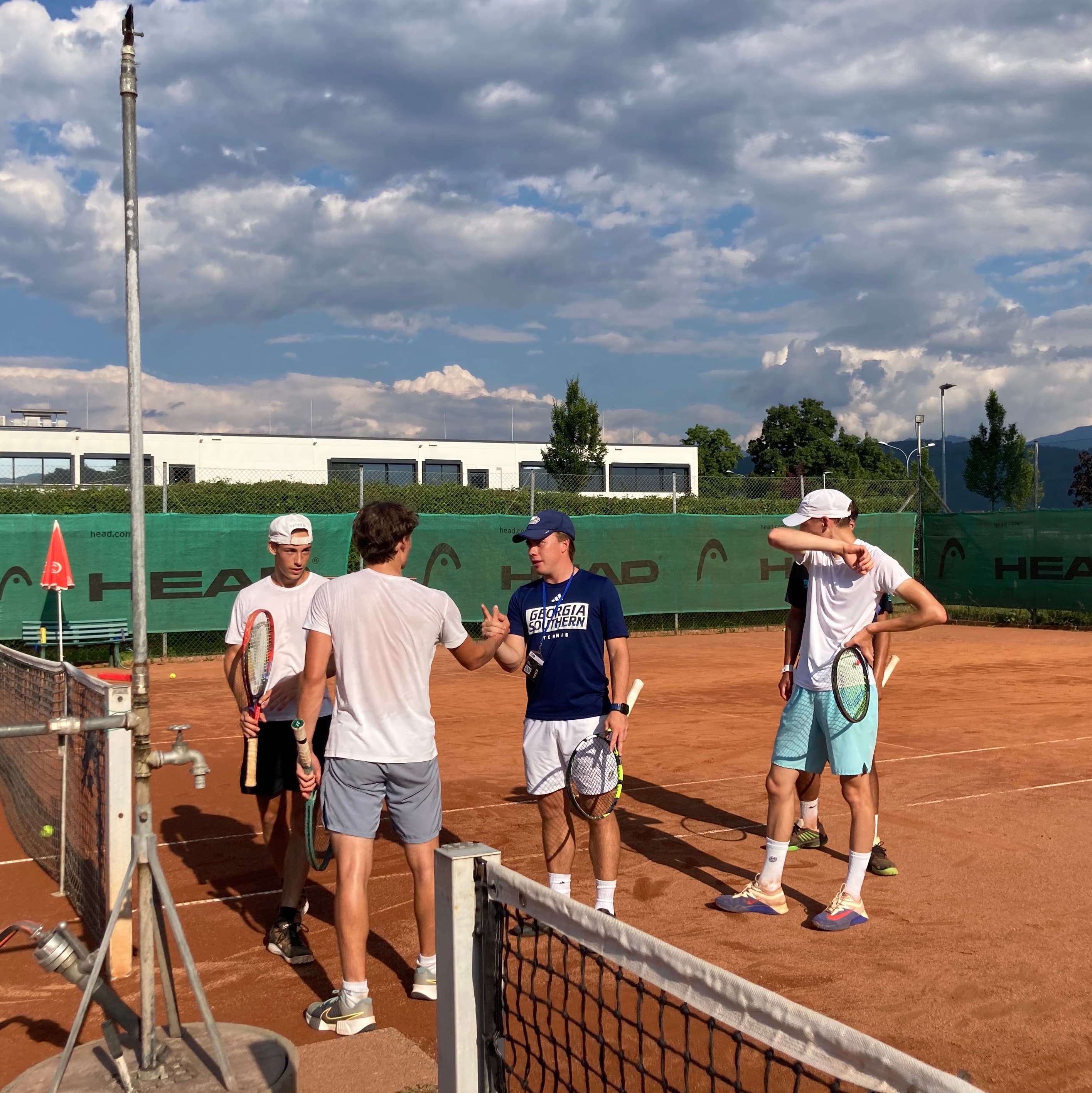 College Tennis Coach Greeting Student-Athlete A college tennis coach in a navy shirt shaking hands with a student-athlete on a clay court, surrounded by more tennis players.